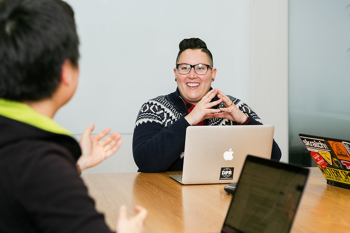 A gender fluid woman smiling while working on her mac and collaborating with a coworker